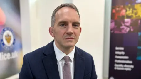 A man with short grey hair in a navy suit sat on a chair in a meeting room. The background is blurred out however banners advertising West Midlands Police force are still visible in the background.