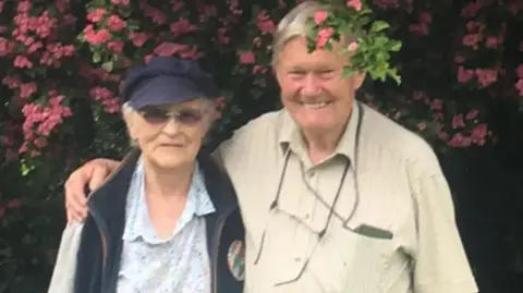 Family photo Ann with her husband Nicholas in front of a flowery hedge