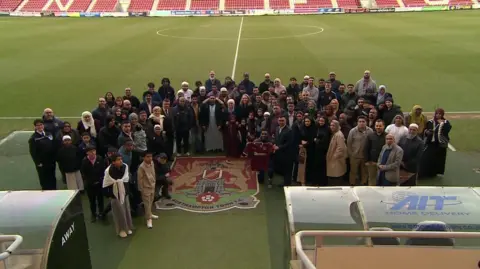 Kate Bradbrook/BBC A large group of people stand by the side of a football pitch.