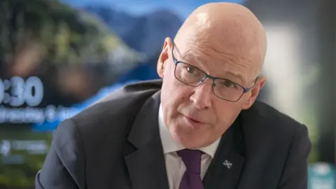 A bald man with glasses - wearing a dark suit, white shirt and purple tie - looks towards the camera while in a seated, hunched position. 