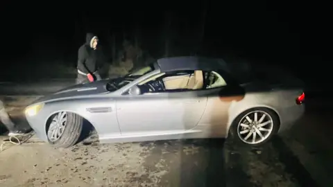 A silver car is by the side of a road with the front left wheel hanging off. It is night time.