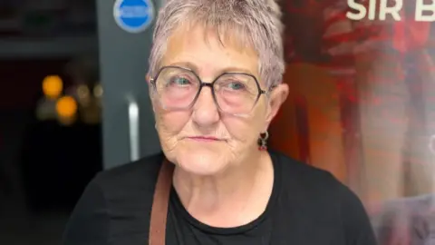 Josie Andrews with short grey hair wearing hexagon shaped glasses and a black t-shirt. She looks emotional after paring tribute to Billy Boston.
