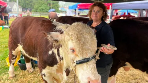 BBC A woman with redish, brown hair standing next to a brown and white cow