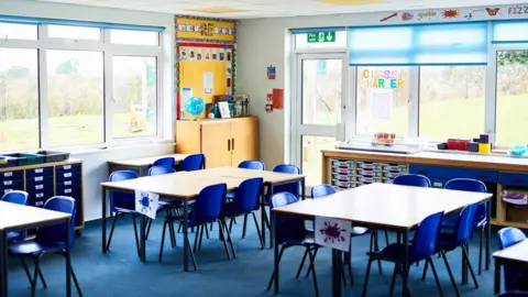 An empty classroom at a primary school in Godalming in Surrey. Blue chairs are placed around various tables. In the background are windows, through which grass can be seen. There are also cupboards, including space for trays.
