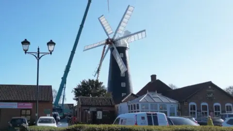 A large traditional windmill stands in the centre of the scene as a crane works beside it. Surrounding buildings, parked vehicles, and a clear blue sky frame the setting.