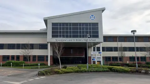 The front of a modern school building. It has a contemporary design with a mix of light-coloured panels and red‑brick sections. Large windows line both the ground and upper floors, creating a bright, open look. The entrance has an overhanging upper section supported by pillars. Above the entrance is a wide set of windows arranged in a grid pattern and under these windows, a sign reads “St Andrew’s and St Bride’s High School.”
