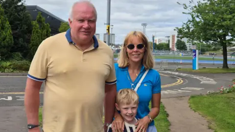 Three people standing outside Scotstoun Stadium - Liffy McIntosh is wearing sunglasses, a blue top and has blonde hair, John McIntosh has a beige top and thinning grey hair, and Harvey McIntosh is a young boy with blond hair and a black and white top on.