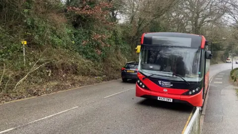 BBC A red bus with a Transport for Cornwall logo on the front drives down a damp Cornish road with trees and hedges on one side and a pavement on the other.