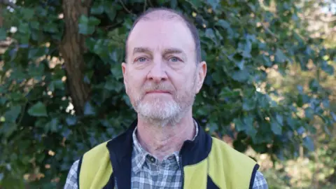 Green Party Martin Fodor wearing a green and white checked shirt underneath a yellow and black gilet. He has short dark hair, green-blue eyes and a white beard, and is looking at the camera with a blank expression. There are trees in the background.