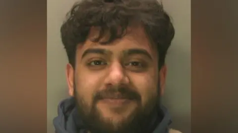 A close-up custody image of a man facing the camera, with short dark hair and a full beard, wearing a dark top against a plain light-coloured background.