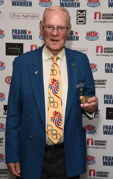 Getty Images A man in his early 80s holds Olympic medals at a boxing dinner event. He is wearing a blue jacket and a yellow tie with the Olympic rings logo. 
