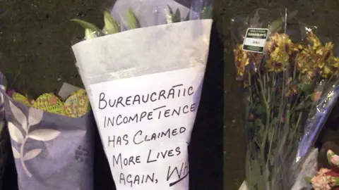 PA Media A message criticising "bureaucratic incompetence" left on a floral tribute following the crash