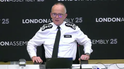 London Assembly Sir Mark Rowley in police uniform sat at a desk at a meeting of the London Assembly's police and crime committee, a laptop and a glass of water in front of him