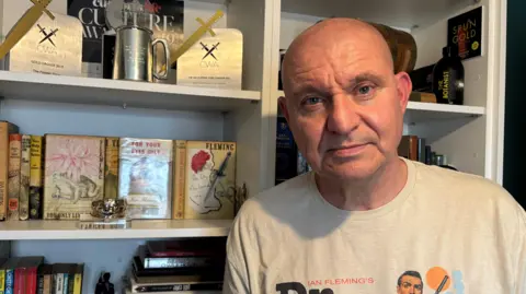 Mike Craven is standing in front of his bookcase and looking at the camera. He is bald and is wearing a 'Ian Fleming's Dr. No' t-shirt. Two gold dagger awards are displayed, along with some of Fleming's books.