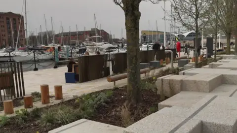 The photo shows concrete seating blocks and new planting along the walkway. Boats and buildings can be seen in the background.