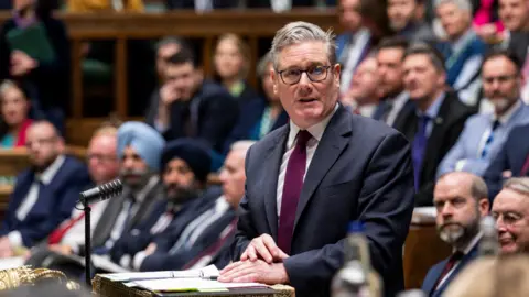 House of Commons Keir Starmer standing in the House of Commons, while his MPs look on. He is wearing a dark suit and a purple tie. 
