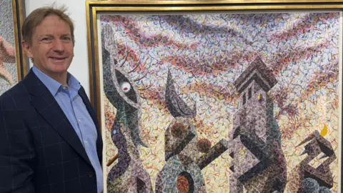 Winsor Birch Grant Ford smiles looking into the camera while standing next to a large surrealist painting, featuring towers and an eye
