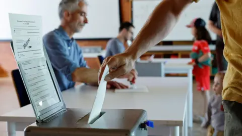 A voter's arm and hand is seen putting a ballot paper into a box, with two officials both wearing blue tops seen sat desks, a girl wearing red shorts and T-shirt and a baseball cap at the far end of the second of the desks