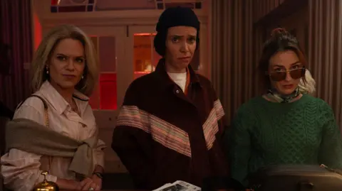 @Netflix,Inc. Three women.
On the left one has shoulder length blonde hair and is wearing a striped blouse with a jumper over her shoulders.
In the middle is a women with brown bobbed hair, She is wearing a beanie hat
On the right is a women with dark hair pulled back into a ponytail, She is wearing a green knitted jumper, a scarf around her neck and sunglasses.