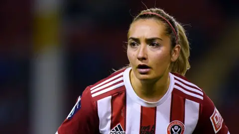 A head-and-shoulders photo of Maddy Cusack. She has a focused expression, appears to be mid-game, and is looking across the image to the left. She is wearing the red and white Sheffield United kit. She has her brown-blonde hair in a high ponytail and is wearing a pink alice band.