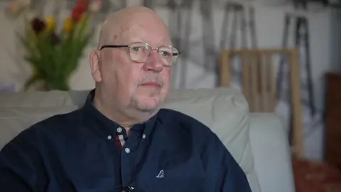 John Kiely sitting on a sofa in his room. He is bald and wears glasses and a long-sleeved navy blue shirt.