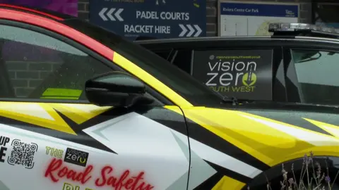 A close-up of a vehicle branded for the Vision Zero South West road safety initiative. The car features a bold, colourful design, with sharp yellow, white, and black geometric patterns and red accents. The text "Road Safety" and "Vision Zero" is prominently displayed.