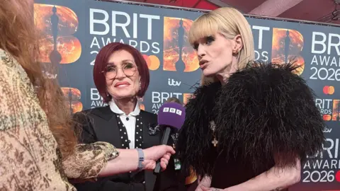 A woman with red hair and a woman with blonde hair and a fur coat, stand on a red carpet speaking into a microphone.