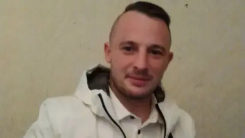 Police Scotland Picture of a man with black hair, shaved at the sides. He has heavy stubble and is half smiling. He is wearing a white jacket with a black lined hood and a white shirt with black buttons.