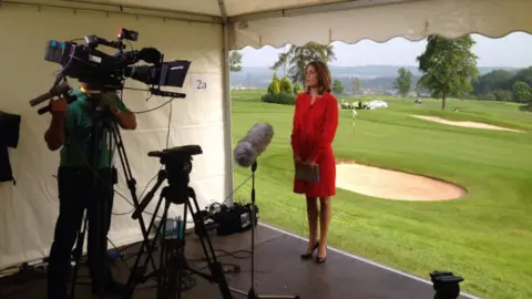 Andrea Byrne / Y Lolfa Andrea Byrne mengenakan gaun merah berdiri di depan kamera dan mikrofon siap menyajikan berita ITV Wales