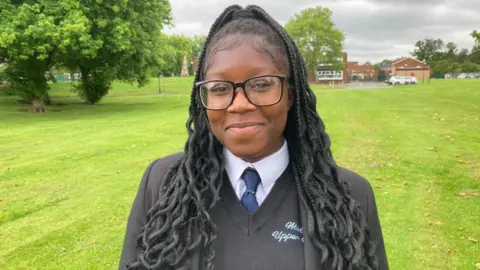 Alex Pope/BBC Franklina is photographed on a green field outside the school building, she is wearing glasses and a dark school uniform, with a blue tie