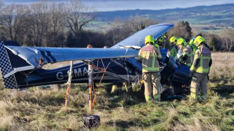 Plane wreckage in a field. Firefighters stand on the right of the craft