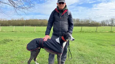 Vikki Irwin/BBC A man is standing on grass next to a fence with sheep in the background. He is holding a lead with a greyhound attached to it. The dog is wearing a blue jacket with red trim and a red collar. The man is wearing a navy jacket with a red lining showing. He is also wearing a grey cap and dark glasses.