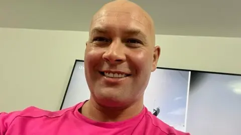 Head and shoulders image of Kirk Marsden, He is bald, smiling and wearing a bright pink tshirt