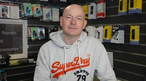 A man wearing a grey hooded jumper smiles at the camera while standing in a shop with electronic products hung up on the wall behind him.