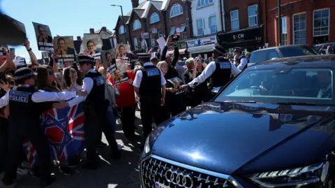 A black Audi is driven and protesters stand beside holding signs reading 'Keir Starmer Jew harmer'