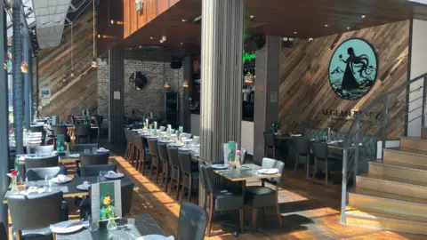 A modern restaurant dining area with neatly arranged tables and chairs and sunlight streaming in through large windows.
