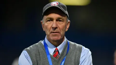 Martin St Quinton standing with his arms folded, wearing a blue shirt and grey jumper and a cap with Gloucester's badge