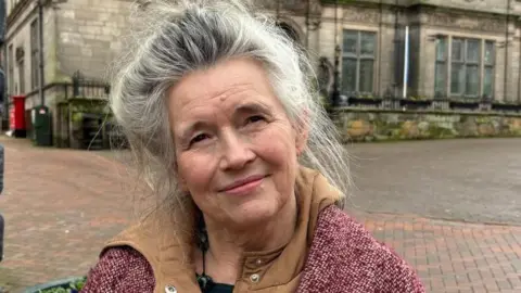 Charlie Stubbs/BBC News A woman with big grey hair, a purple jacket with a chocolate-brown interior. She is stood in a courtyard with a grey building with black framed windows in the background.