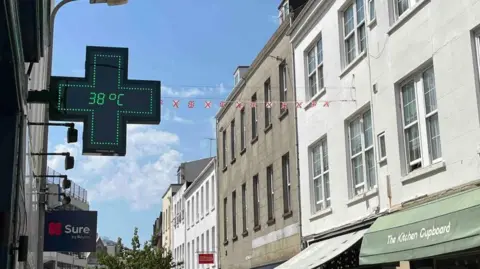 A pharmacy sign, in a plus shape, shows green L E D lights, with the temperature reading 38 degrees celsius. It's on a high street, with white and grey 3 storey buildings on the right hand. A shop veranda on one is green and says 'The Kitchen Cupboard'. Bunting hung across the street shows the Jersey and British flags in front of a blue sky.
