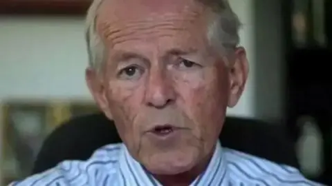 John Smyth, pictured talking to a camera before his death in 2018, wears a white striped shirt