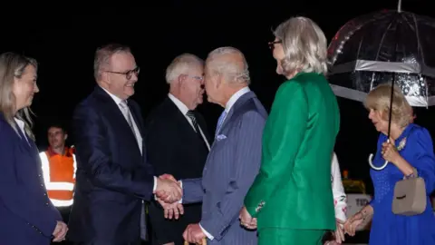 Brooke Mitchell / Getty Images Een glimlachende Anthony Albanese staat in de rij en begroet koning Charles en koningin Camilla als ze aankomen op de internationale luchthaven van Sydney 