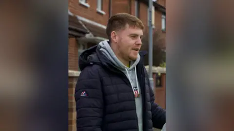 Ronan McLaughlin A man is standing on a street with terraced houses behind him. He has short brown hair and is wearing a black coat with a grey hoodie.