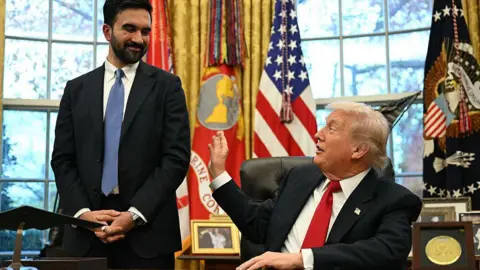 Getty Images Mamdani é visto ao lado de Trump, que está sentado atrás de sua mesa no Salão Oval. Ambos riem e Trump, brincando, estende a mão para dar um tapinha no braço dele