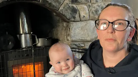 Nicola Graham, who is wearing a black hoodie and black-rimmed glasses and has a brown ponytail, sits next to a black woodburning stove with a chrome chimney. She is holding her daughter Neavh, an 8-month-old baby wearing a white babygrow