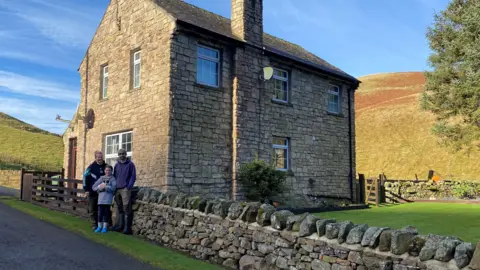 Harry and Megan Byatt are standing with a child outside a modern, two-storey stone farmhouse next to a single track road. Behind them the Cheviot hills and the house are bathed in late evening sunshine 
