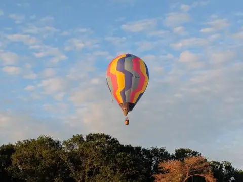 BBC Weather Watchers/SpannaH A multi-coloured hot-air balloon hangs in the air over a green forest.