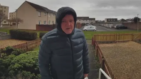 A man in a hooded jacket on the front doorstep of a house. Several other homes and cars are visible in the background.