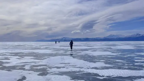 Anya Eames A huge frozen lake, with some snow in patches across it. There are tall snow-capped mountains in the distance, and four people can be seen ahead of her running on the lake. There are some clouds drifting across the blue skies. 