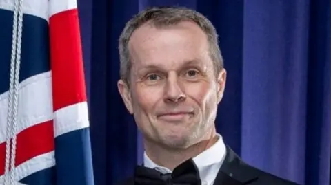 West Midlands Police Tim Rogers, a man wearing a white shirt with a black bow-tie and black suit jacket, is stood against a navy blue backdrop with a Union Jack flag to the left hand side of the image.
