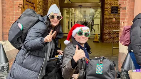 Fran Ackroyd Steve Ackroyd in a wheelchair wearing a Santa hat and festive glasses, alongside his wife Fran, both smiling and posing for the camera.